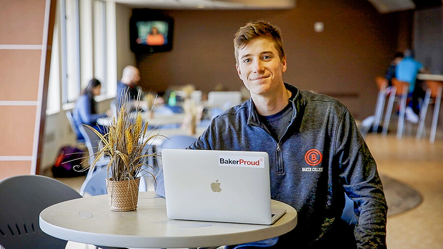 A student sits at a table with a laptop in a residence hall common area. The laptop has a "BakerProud" sticker.