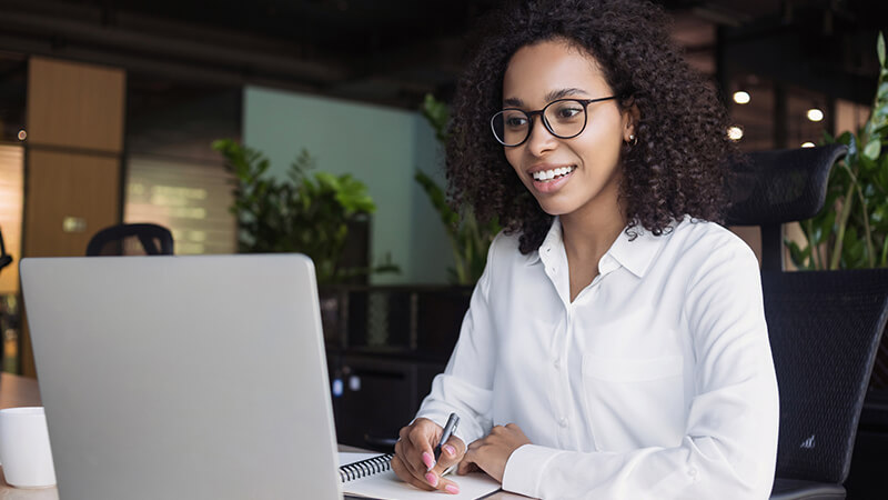 A woman sits in front of a laptop and takes notes in a notebook