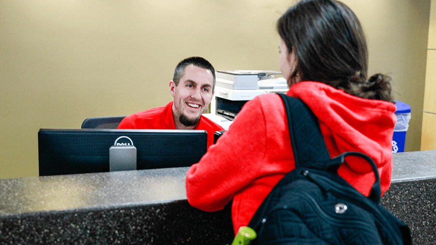 A staff member in a red T-shirt assists a student with a technology question.